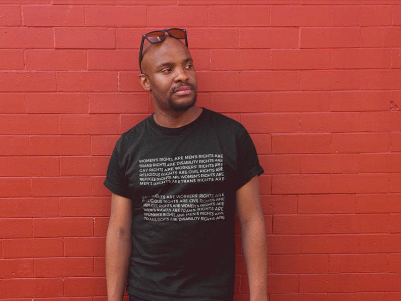 Puede incluir: Una persona lleva una camiseta negra con texto blanco que dice "Los derechos de las mujeres son los derechos de los hombres, los derechos trans son los derechos de las personas con discapacidad, los derechos de los homosexuales son los derechos de los trabajadores, los derechos religiosos son los derechos civiles, los derechos de los refugiados son los derechos de las mujeres, los derechos de los hombres son los derechos trans". La persona est&aacute; de pie frente a una pared de ladrillo rojo.