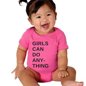 Pu&ograve; includere: Un body rosa per neonato con testo nero che recita "GIRLS CAN DO ANY-THING".