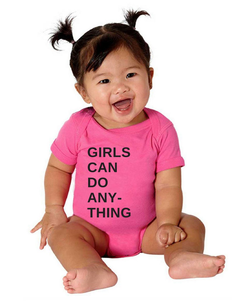 Pu&ograve; includere: Un body rosa per neonato con testo nero che recita "GIRLS CAN DO ANY-THING".