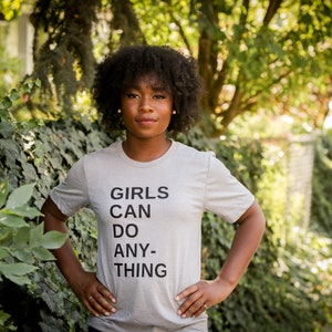 May include: A woman with dark curly hair is wearing a grey t-shirt with the text "GIRLS CAN DO ANY-THING" printed on it.
