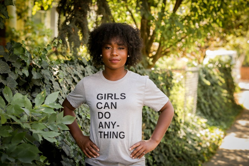 May include: A woman with dark curly hair is wearing a grey t-shirt with the text "GIRLS CAN DO ANY-THING" printed on it.