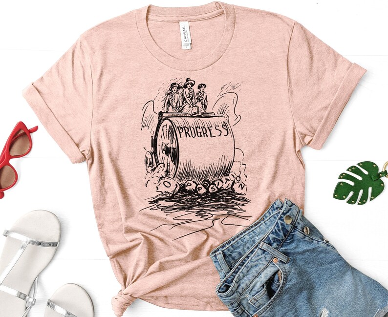 May include: A pink t-shirt with a black and white illustration of a steamroller with the word "PROGRESS" on it.  Four people are riding on the steamroller.