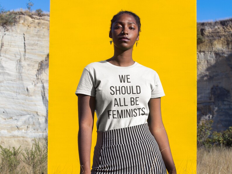 Puede incluir: Una persona lleva una camiseta blanca con el texto "WE SHOULD ALL BE FEMINISTS" impreso en ella. La persona est&aacute; de pie frente a un fondo amarillo.