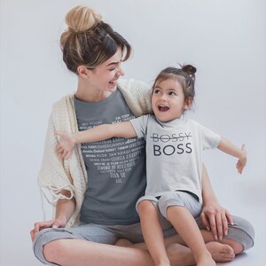 May include: A woman and a young girl are sitting on the floor, smiling at each other. The woman is wearing a grey t-shirt with a list of women's names printed on it. The girl is wearing a white t-shirt with the words "Bossy Boss" printed on it.