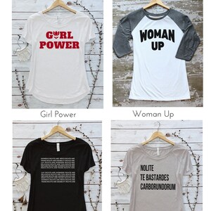 May include: Four different t-shirt designs with various slogans. The first shirt is white with red text that says "Girl Power". The second shirt is white with a gray sleeve and black text that says "Woman Up". The third shirt is black with white text that says "Women's Rights are Men's Rights are Trans Rights are Disability Rights are Gay Rights are Workers Rights are Religious Rights are Civil Rights are Refugee Rights are Women's Rights are Men's Rights are Trans Rights are". The fourth shirt is gray with black text that says "Nolite Te Bastardes Carborundorum".