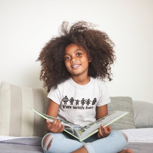May include: A young person with curly hair is sitting on a bed and reading a book. They are wearing a white t-shirt with a black graphic that says "I'm with her." and a pair of light blue jeans.