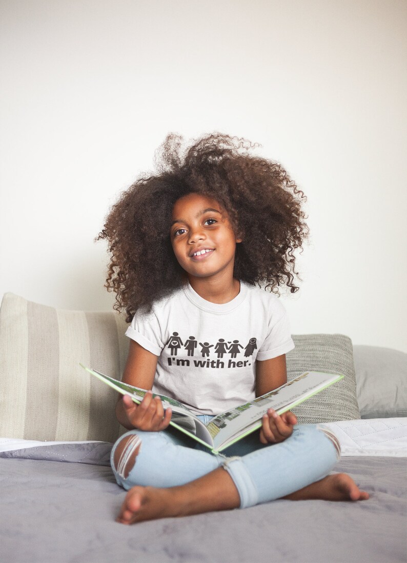 May include: A young person with curly hair is sitting on a bed and reading a book. They are wearing a white t-shirt with a black graphic that says "I'm with her." and a pair of light blue jeans.