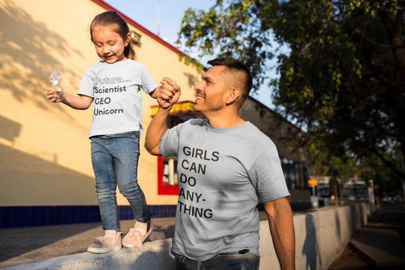 Puede incluir: Un padre y una hija llevan camisetas grises claras a juego. La camiseta de la hija dice "Future... Scientist CEO Unicorn". La camiseta del padre dice "Girls Can Do Anything".
