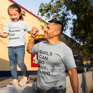 May include: A father and daughter wearing matching light gray t-shirts. The daughter's shirt says "Future... Scientist CEO Unicorn". The father's shirt says "Girls Can Do Anything".