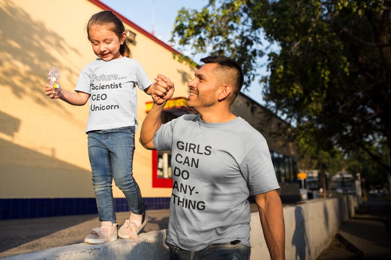 May include: A father and daughter wearing matching light gray t-shirts. The daughter's shirt says "Future... Scientist CEO Unicorn". The father's shirt says "Girls Can Do Anything".