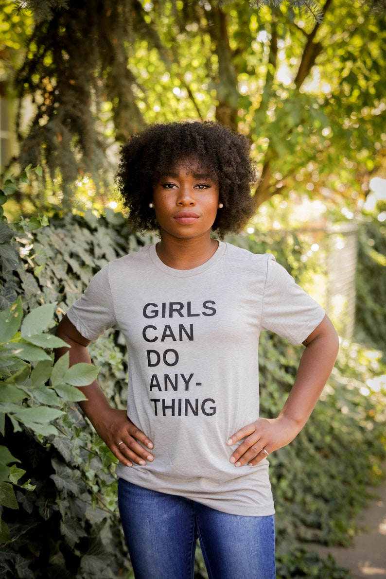 Puede incluir: Una persona lleva una camiseta gris con el texto "GIRLS CAN DO ANY-THING" impreso en ella. Est&aacute; de pie frente a un fondo verde frondoso.