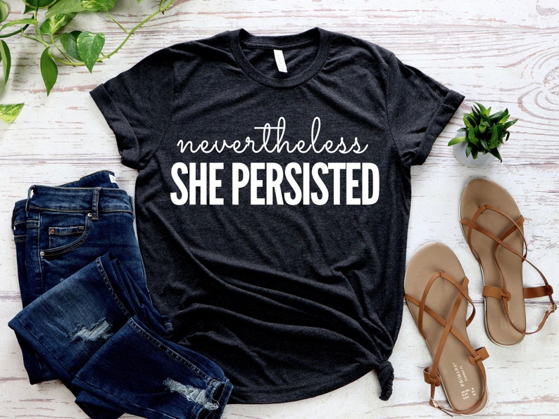 May include: A dark grey t-shirt with the text "nevertheless SHE PERSISTED" printed in white.
