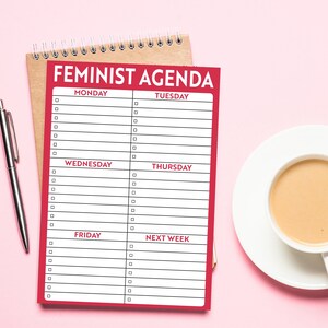 K&ouml;nnte beinhalten: Ein roter Planer mit dem Aufdruck "Feminist Agenda" auf der Vorderseite. Der Planer enth&auml;lt eine Liste der Wochentage mit leeren Zeilen zum Schreiben.