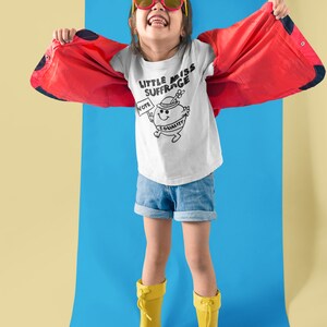 May include: A young person wearing a white t-shirt with a black and white illustration of a cartoon character holding a sign that says "Vote" and a banner that says "Equality". The t-shirt also has the text "Little Miss Suffrage". The person is wearing red sunglasses, blue denim shorts, and yellow rain boots. The person is standing on a blue surface with a yellow surface in the background.