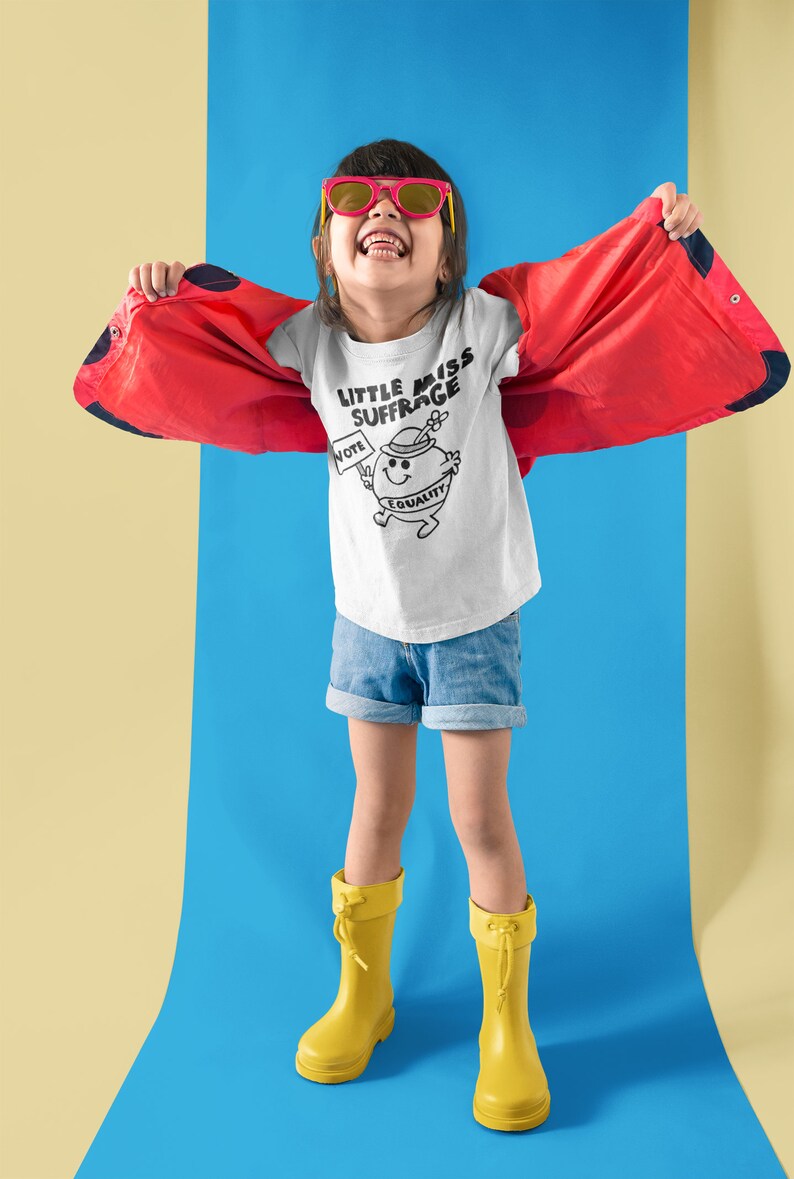 May include: A young person wearing a white t-shirt with a black and white illustration of a cartoon character holding a sign that says "Vote" and a banner that says "Equality". The t-shirt also has the text "Little Miss Suffrage". The person is wearing red sunglasses, blue denim shorts, and yellow rain boots. The person is standing on a blue surface with a yellow surface in the background.