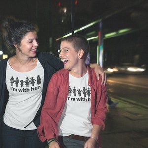 Pu&ograve; includere: Due persone indossano magliette bianche con una grafica di figure stilizzate che si tengono per mano e il testo "I'm with her." La persona a sinistra indossa un cardigan rosso. Sorridono e si guardano.