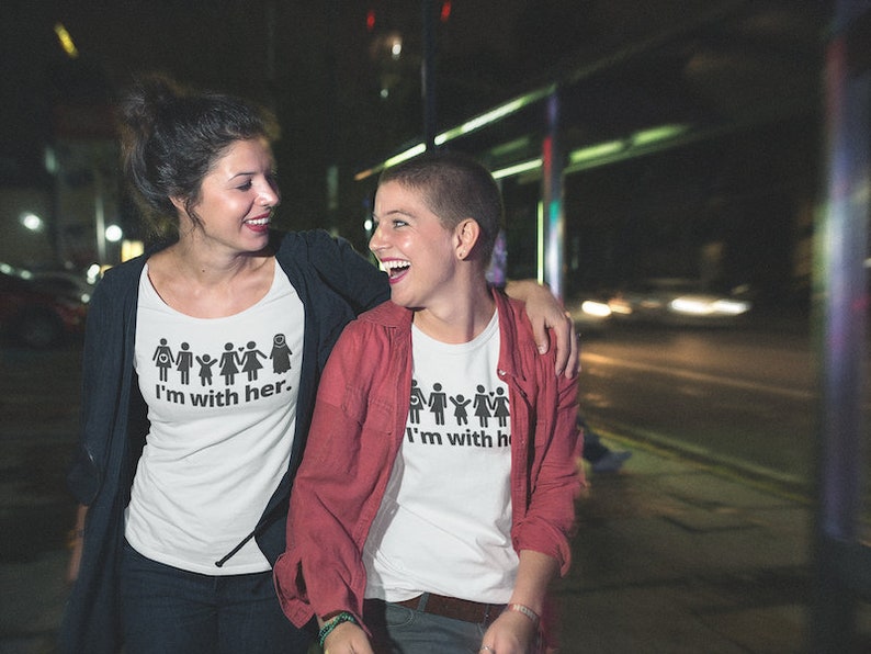 Pu&ograve; includere: Due persone indossano magliette bianche con una grafica di figure stilizzate che si tengono per mano e il testo "I'm with her." La persona a sinistra indossa un cardigan rosso. Sorridono e si guardano.