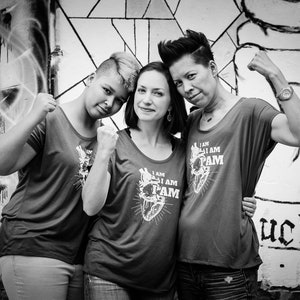 Pu&ograve; includere: Tre persone stanno davanti a un muro con graffiti, ciascuna con una maglietta grigia con il testo "I AM I AM PAM" e un grafico di un cuore. La persona a sinistra indossa i jeans, la persona al centro indossa i jeans e la persona a destra indossa pantaloncini neri.