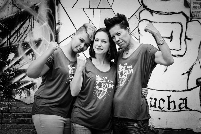 Pu&ograve; includere: Tre persone stanno davanti a un muro con graffiti, ciascuna con una maglietta grigia con il testo "I AM I AM PAM" e un grafico di un cuore. La persona a sinistra indossa i jeans, la persona al centro indossa i jeans e la persona a destra indossa pantaloncini neri.