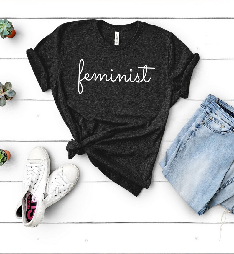 May include: A dark gray t-shirt with the word "feminist" written in white cursive script.