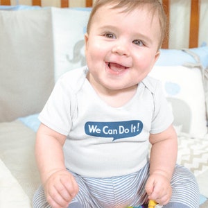 Pu&ograve; includere: Un bambino piccolo che indossa un body bianco con strisce blu e il testo "We Can Do It!" in un fumetto. Il bambino &egrave; seduto su una coperta a motivi bianchi e grigi.