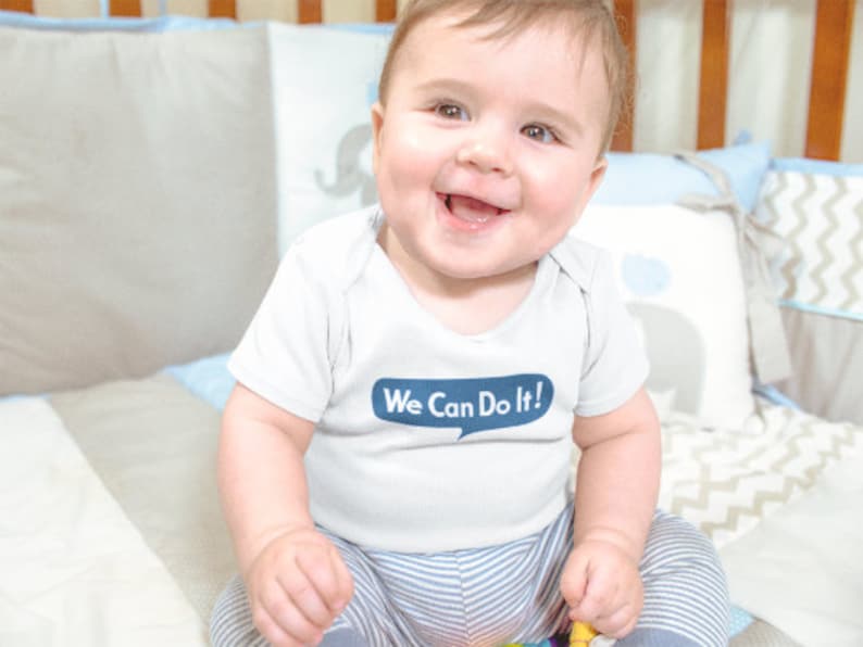 Pu&ograve; includere: Un bambino piccolo che indossa un body bianco con strisce blu e il testo "We Can Do It!" in un fumetto. Il bambino &egrave; seduto su una coperta a motivi bianchi e grigi.
