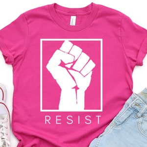 May include: Pink t-shirt with a white raised fist graphic and the word "RESIST" printed below.