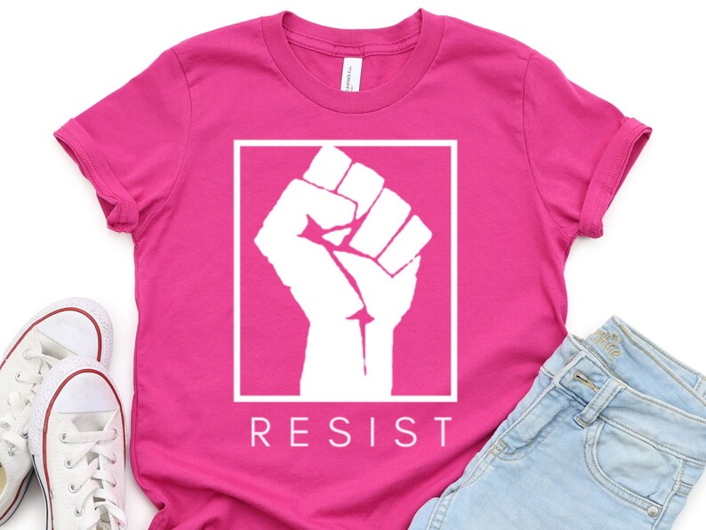 May include: Pink t-shirt with a white raised fist graphic and the word "RESIST" printed below.