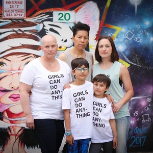 Pu&ograve; includere: Un gruppo di persone sta in piedi davanti a un muro di graffiti colorato. Il muro presenta una grande tigre bianca con accenti rosa e blu. Le persone indossano magliette bianche con la scritta "Girls Can Do Anything" stampata su di esse. Due delle persone sono bambini.