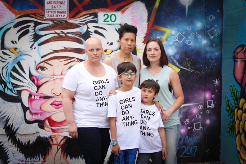 Pu&ograve; includere: Un gruppo di persone sta in piedi davanti a un muro di graffiti colorato. Il muro presenta una grande tigre bianca con accenti rosa e blu. Le persone indossano magliette bianche con la scritta "Girls Can Do Anything" stampata su di esse. Due delle persone sono bambini.
