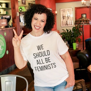 May include: A woman wearing a white t-shirt with the text "WE SHOULD ALL BE FEMINISTS" in black lettering. She is smiling and giving a peace sign with her left hand. She is wearing blue jeans and is standing in a cafe.