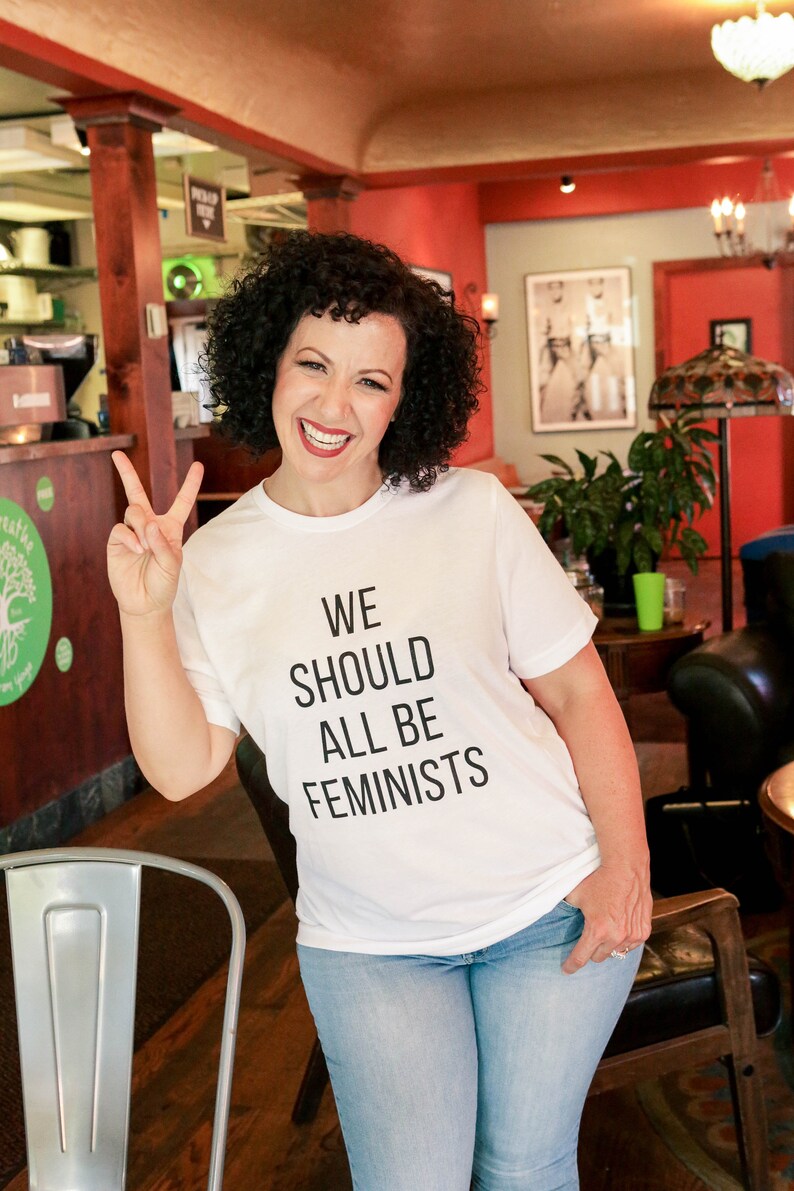 May include: A woman wearing a white t-shirt with the text "WE SHOULD ALL BE FEMINISTS" in black lettering. She is smiling and giving a peace sign with her left hand. She is wearing blue jeans and is standing in a cafe.