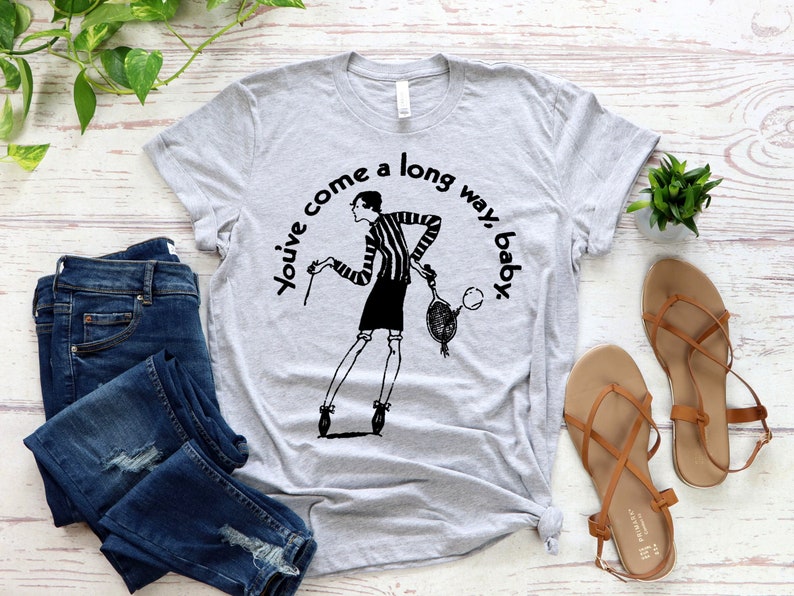 Puede incluir: Camiseta gris con una ilustraci&oacute;n en blanco y negro de una mujer sosteniendo una raqueta de tenis. El texto "You've come a long way, baby" est&aacute; impreso en la camiseta.