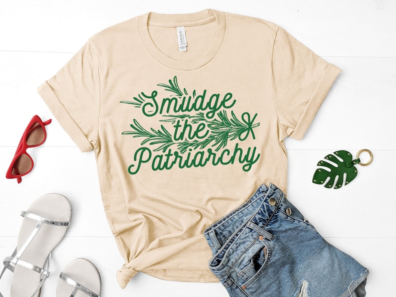 May include: A beige t-shirt with a green leafy design and the text "Smudge the Patriarchy".