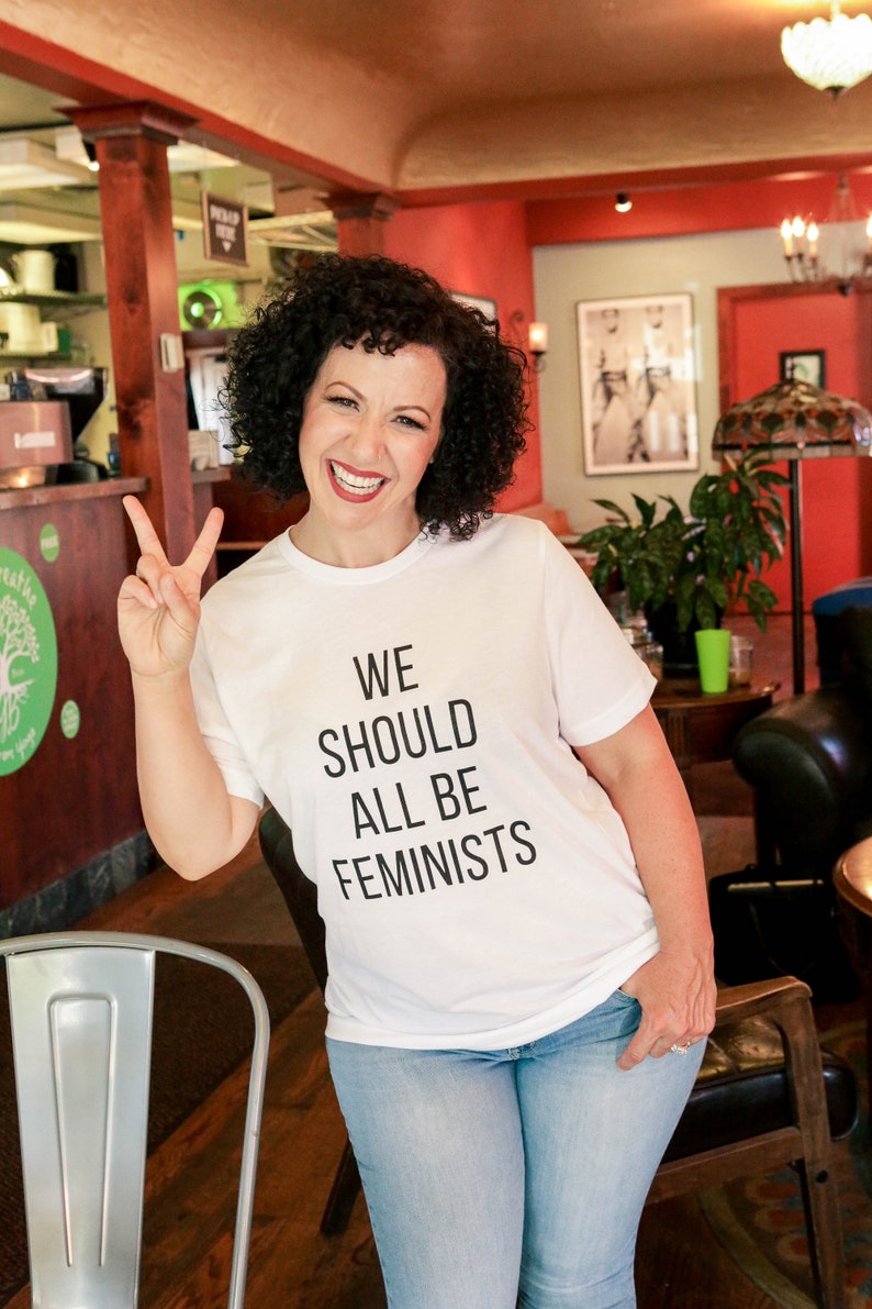 Puede incluir: Una mujer con una camiseta blanca con el texto "WE SHOULD ALL BE FEMINISTS" en letras negras. Est&aacute; sonriendo y haciendo un signo de paz con su mano izquierda. Lleva pantalones vaqueros azules y est&aacute; de pie en una cafeter&iacute;a.