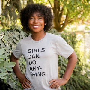 May include: A woman wearing a grey t-shirt with the text "GIRLS CAN DO ANY-THING" printed on it.