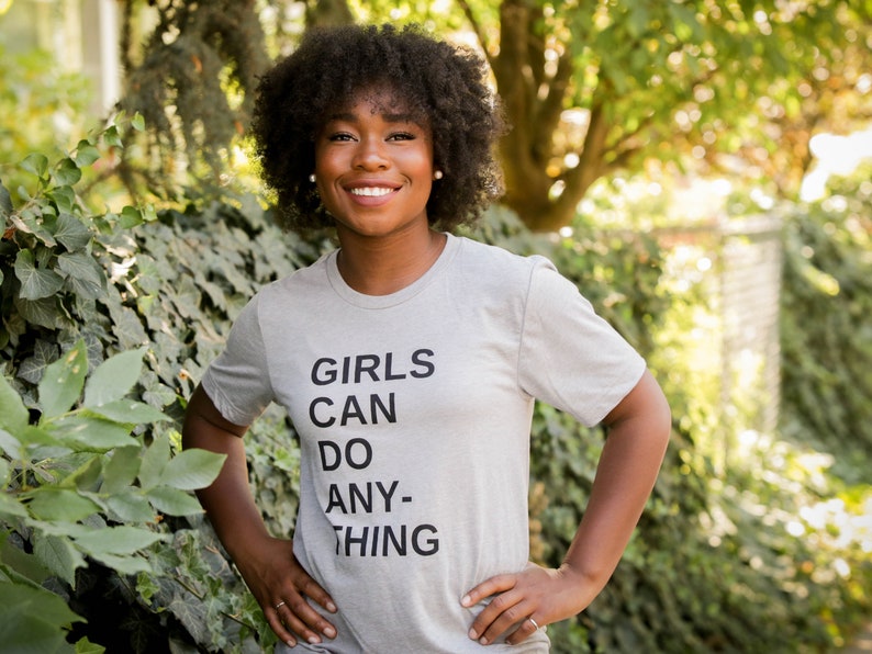 May include: A woman wearing a grey t-shirt with the text "GIRLS CAN DO ANY-THING" printed on it.