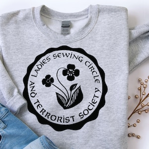 May include: Gray sweatshirt with a black circle graphic that says "And Ladies Sewing Circle Society" with a stylized flower design.