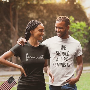 May include: A couple wearing matching t-shirts with feminist slogans. The woman's shirt says "feminist" in a cursive font. The man's shirt says "We should all be feminists".