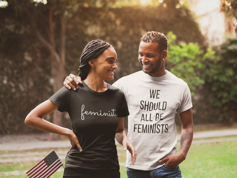 May include: A couple wearing matching t-shirts with feminist slogans. The woman's shirt says "feminist" in a cursive font. The man's shirt says "We should all be feminists".