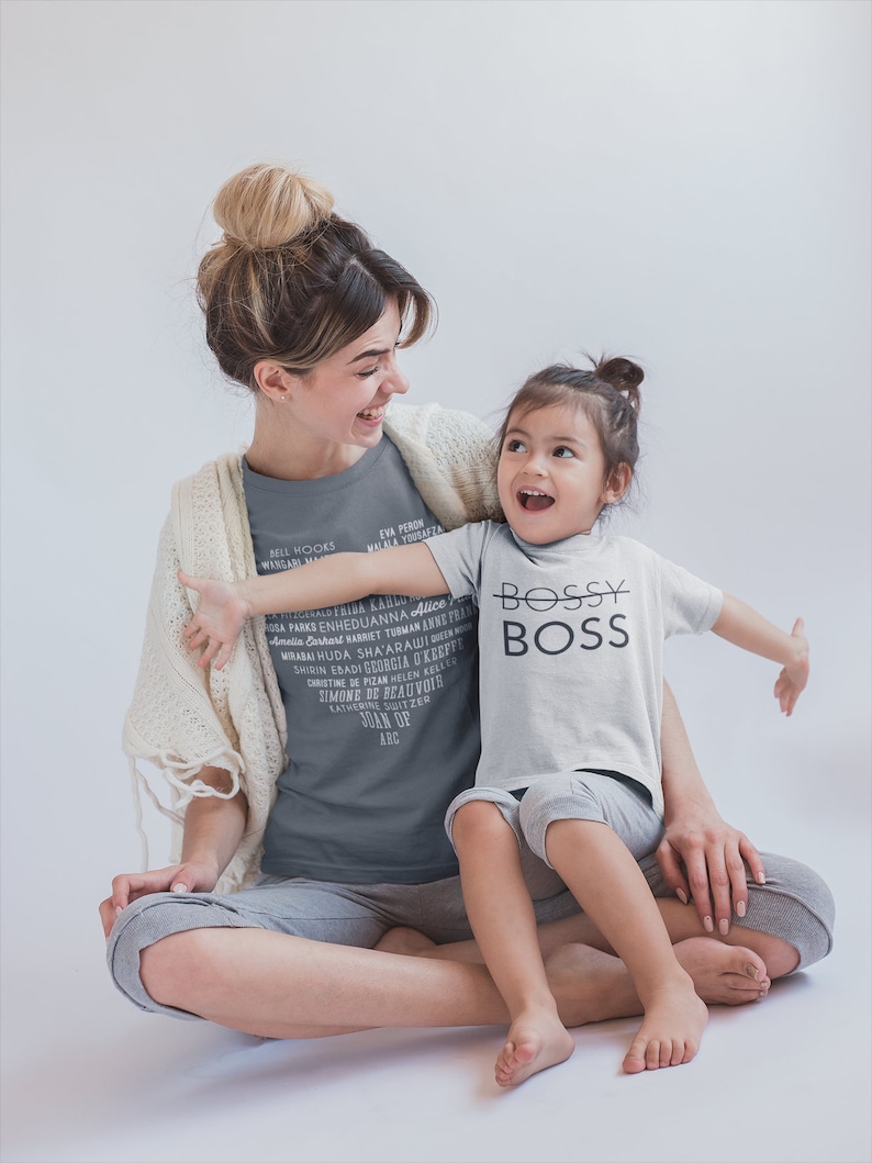 Puede incluir: Una mujer y una ni&ntilde;a peque&ntilde;a est&aacute;n sentadas juntas. La mujer lleva una camiseta gris con una lista de nombres de mujeres impresos en ella. La ni&ntilde;a lleva una camiseta blanca con las palabras "Bossy Boss" impresas en ella. Ambas est&aacute;n sonriendo y mir&aacute;ndose.