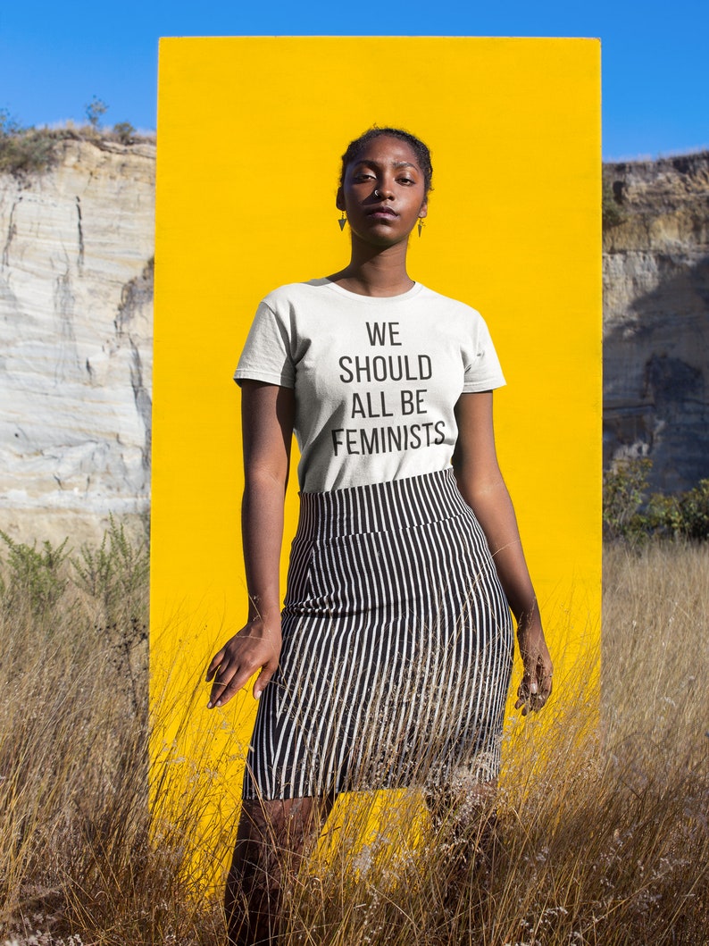 May include: A person wearing a white t-shirt with the text "WE SHOULD ALL BE FEMINISTS" printed on it. The person is standing in front of a yellow background.