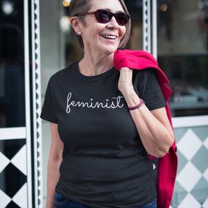 May include: A woman wearing a black t-shirt with the word "feminist" written in white cursive script. She is smiling and wearing sunglasses. She is standing in front of a store with a sign that says "COF&Eacute;".