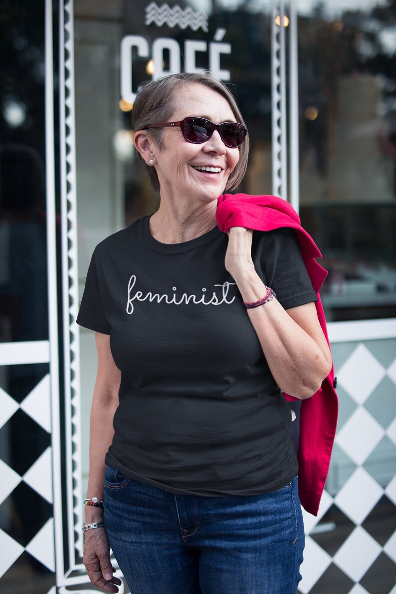 May include: A woman wearing a black t-shirt with the word "feminist" written in white cursive script. She is smiling and wearing sunglasses. She is standing in front of a store with a sign that says "COF&Eacute;".