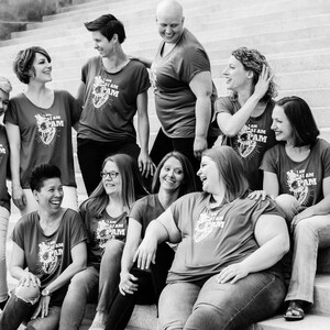 Pu&ograve; includere: Un gruppo di donne indossa magliette rosse con il testo "I AM PAM" e una grafica a forma di cuore. Sono in piedi su una scalinata e sorridono.