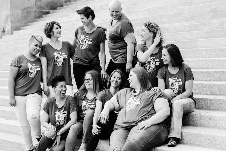 Pu&ograve; includere: Un gruppo di donne indossa magliette rosse con il testo "I AM PAM" e una grafica a forma di cuore. Sono in piedi su una scalinata e sorridono.