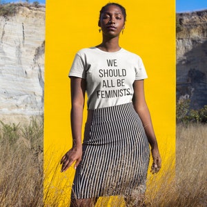 Puede incluir: Una persona lleva una camiseta blanca con el texto "WE SHOULD ALL BE FEMINISTS" impreso en ella. La persona est&aacute; de pie frente a un fondo amarillo.