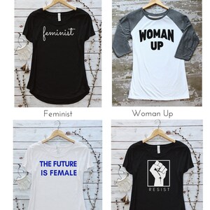May include: Four different t-shirt designs with feminist themes. The shirts are displayed on hangers against a white wood background. The designs include "feminist", "woman up", "the future is female", and "resist" with a raised fist graphic.