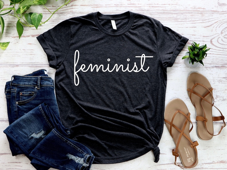 May include: A dark gray t-shirt with the word "feminist" written in white cursive script.
