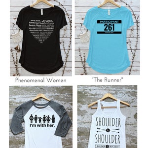 May include: Four different t-shirt designs on hangers. The first shirt is black with white text listing the names of many women. The second shirt is light blue with black text that says "Participant 261 Boston 1967". The third shirt is white with a gray sleeve and black text that says "I'm with her." and has a graphic of a family. The fourth shirt is white with black text that says "Shoulder to Shoulder Bolder & Bolder" and has a graphic of two arrows pointing towards each other.